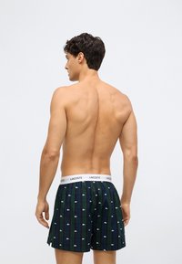 Bărbat văzut din spate, purtând boxeri în carouri verzi și bleumarin, cu un brâu alb cu logo-ul Lacoste și mici modele de crocodili albi.