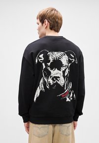 Μαύρη ζακέτα με λευκό γραφικό σχέδιο ενός σκυλιού στην πλάτη, με κόκκινη ετικέτα που γράφει "HUGO". Ραμμένο περιθώριο στον λαιμό και στα μανίκια.