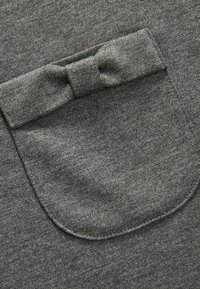 Grijze stof met een gladde textuur, voorzien van een ronde zak versierd met een klein strikdetail. Het materiaal lijkt zacht en gebreid.