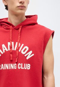 Young person wearing sleeveless red hoodie with "Champion Training Club" text, facing slightly left on plain light background.
