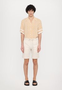 Chemise à manches courtes, orange clair à motifs avec des accents blancs, associée à un short crème. Complétée par des sandales noires. Fond blanc uni.
