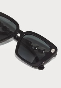 Óculos de sol quadrados pretos com lentes escuras, apresentando um acabamento brilhante, uma armação grossa e um detalhe de cristal de lado.