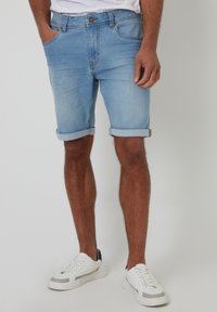 Pantalones cortos de mezclilla azul claro con puños enrollados, diseño de cinco bolsillos y cierre de botón frontal. Llevados con zapatillas blancas.