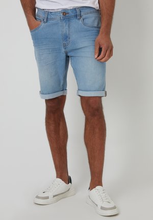 Shorts en denim bleu clair avec revers, design à cinq poches et fermeture à bouton sur le devant. Portés avec des baskets blanches.