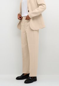 Beiga skräddarsydda byxor i mjukt tyg, med smal passform och raka ben, i kombination med svarta loafers för en kontrasterande look.