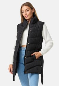 Musta puffervest, jossa on korkea kaulus, tikattu muotoilu, vetoketjusuljenta ja sivutaskut. Päälle pukeutunut valkoisen neuleen ja sinisten farkkujen kanssa.