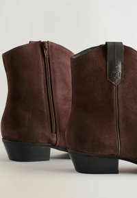 Botas tobilleras de ante marrón con cremallera lateral, una lengüeta de cuero y un acabado ligeramente texturizado con un tacón bajo apilado.