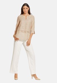 Beige Tunika mit strukturiertem Muster, V-Ausschnitt, Dreiviertelärmel, kombiniert mit weißen weiten Hosen und beigen Absätzen.
