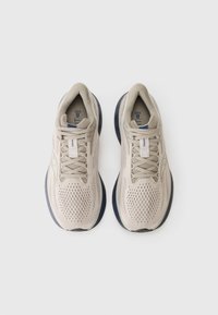 Beige athletic shoes with a mesh upper, round toe, and cushioned navy sole. Features laces and branding on the tongue and side.