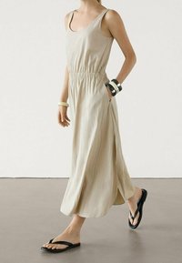 Robe maxi beige sans manches avec une taille élastique, des poches latérales et une silhouette fluide. Portée avec des tongs noires et des bracelets.
