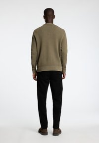 Olīvzaļš adīts džemperis ar teksturētu rakstu, ribotām piedurknēm un apakšmalu, apvienots ar melnām biksēm un brūnām kurpēm, skatīts no aizmugures.