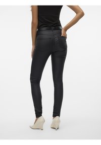 Jean skinny noir en similicuir avec une texture lisse, taille mi-haute et deux poches arrière. Associé à des talons compensés de couleur crème.