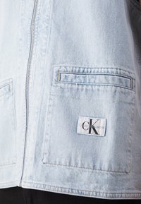 Světle modrá džínová bunda vpředu se zipem, obdélníkovými kapsami a bílou nášivkou s logem Calvin Klein Jeans na spodní kapse.
