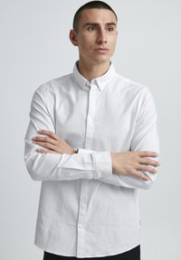 Chemise blanche à manches longues avec col, en tissu légèrement texturé. Le modèle croise les bras sur un fond neutre.