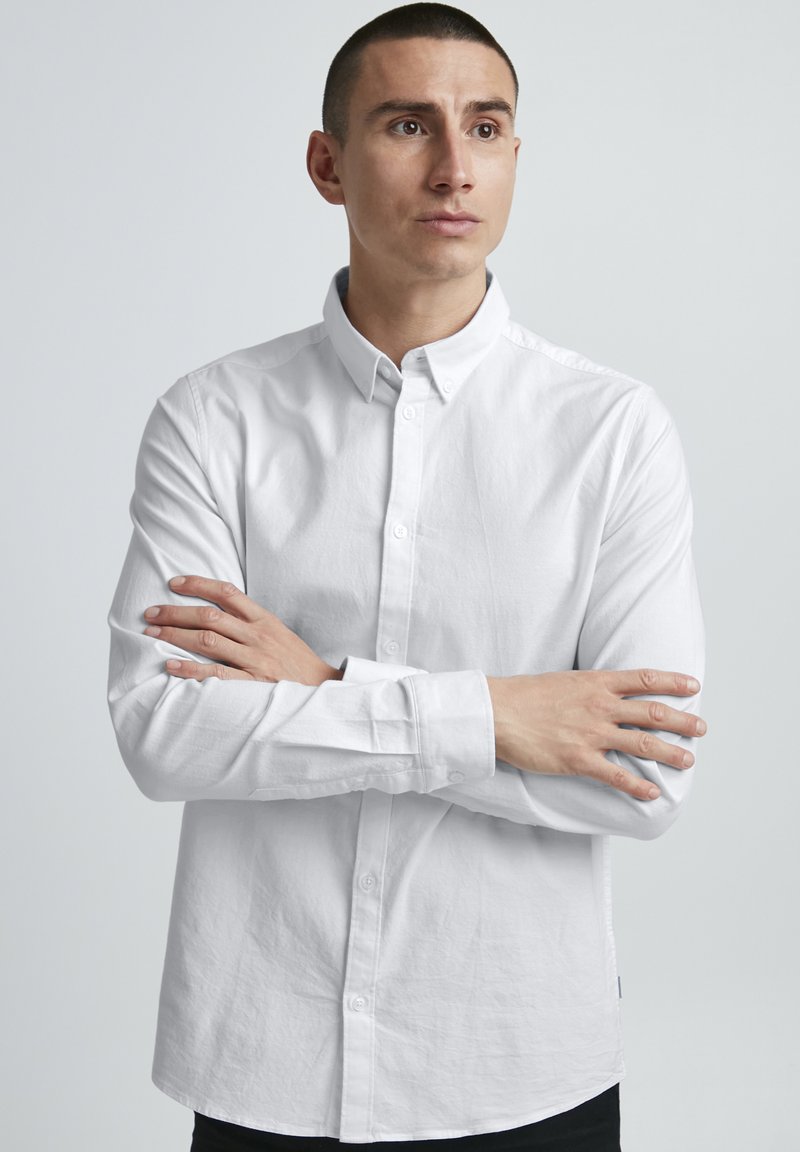 Chemise blanche à manches longues avec col, en tissu légèrement texturé. Le modèle croise les bras sur un fond neutre.