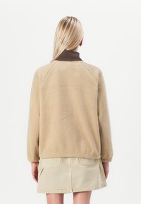 Beige fleece pullover met een bruine kraag, met een zachte textuur, raglanmouwen en een relaxte pasvorm, gedragen over een lichtbeige rok.