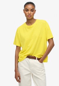 Gul kortærmet t-shirt lavet af blødt stof med en løs pasform og rund halsudskæring, parret med lyse cremefarvede bukser og et fletet brunt bælte.