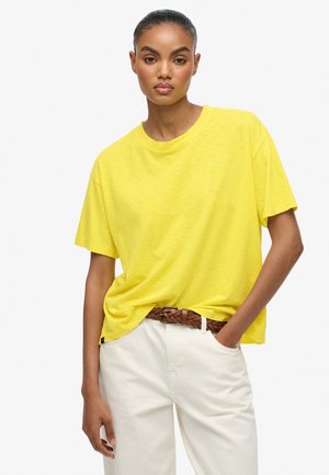 Gul kortærmet t-shirt lavet af blødt stof med en løs pasform og rund halsudskæring, parret med lyse cremefarvede bukser og et fletet brunt bælte.