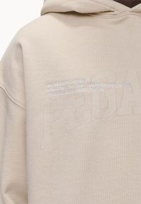 Sweat à capuche beige avec un tissu texturé. L'avant présente le lettrage en relief "PEGA" et un texte brodé en blanc. Capuche attachée, coupe décontractée.