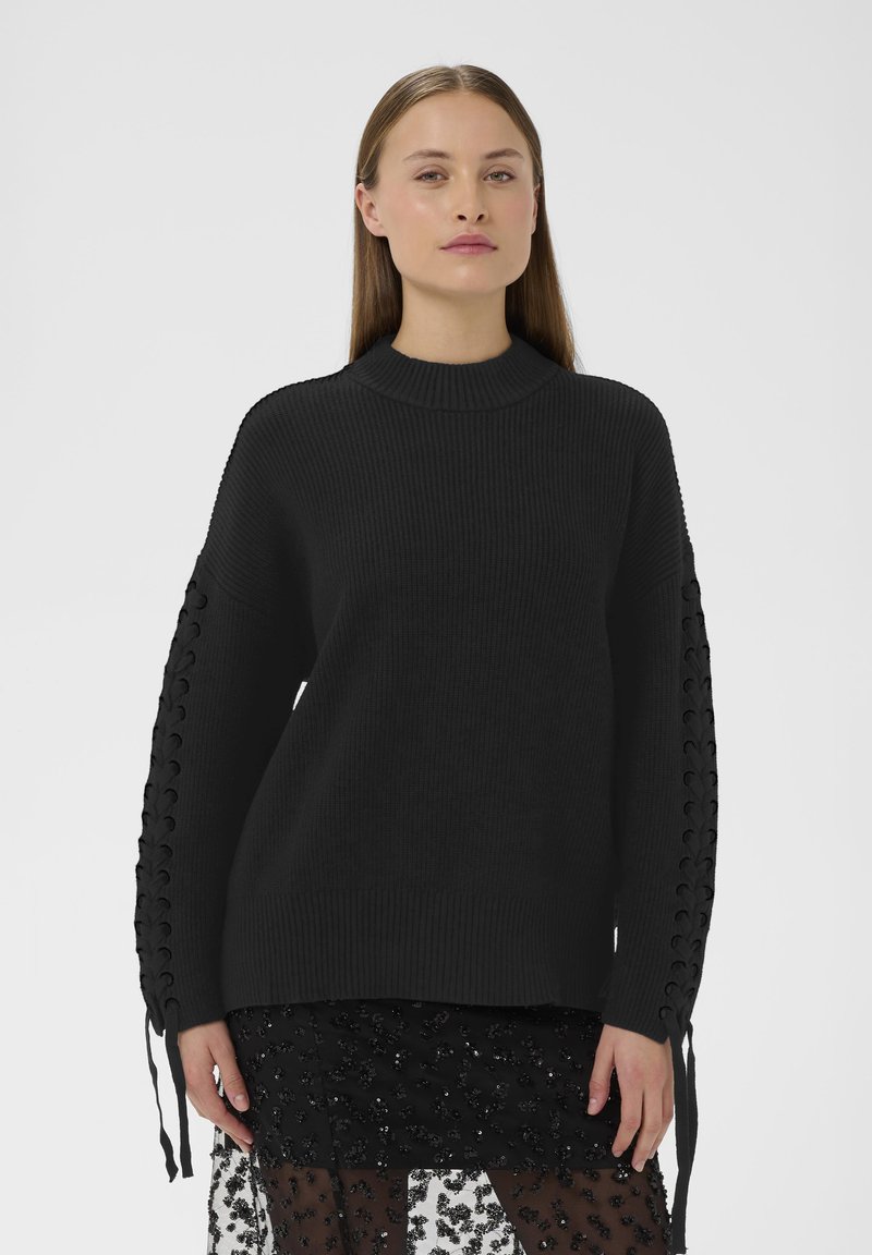Pull en tricot noir avec un col haut et un tissu texturé. Présente des détails à laçage sur les manches, ajoutant un intérêt visuel à la conception.