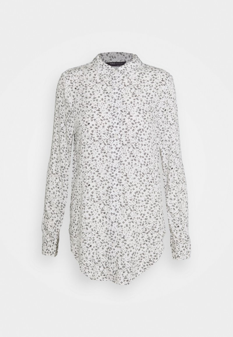 Chemise blanche boutonnée avec manches longues, présentant un motif floral noir, un col pointu et un ourlet arrondi. Texture de tissu douce.