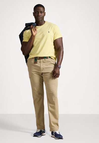 Yellow short-sleeve t-shirt, khaki pants with slight distressing, green and red patterned belt, blue sneakers, and a navy jacket draped over shoulder.