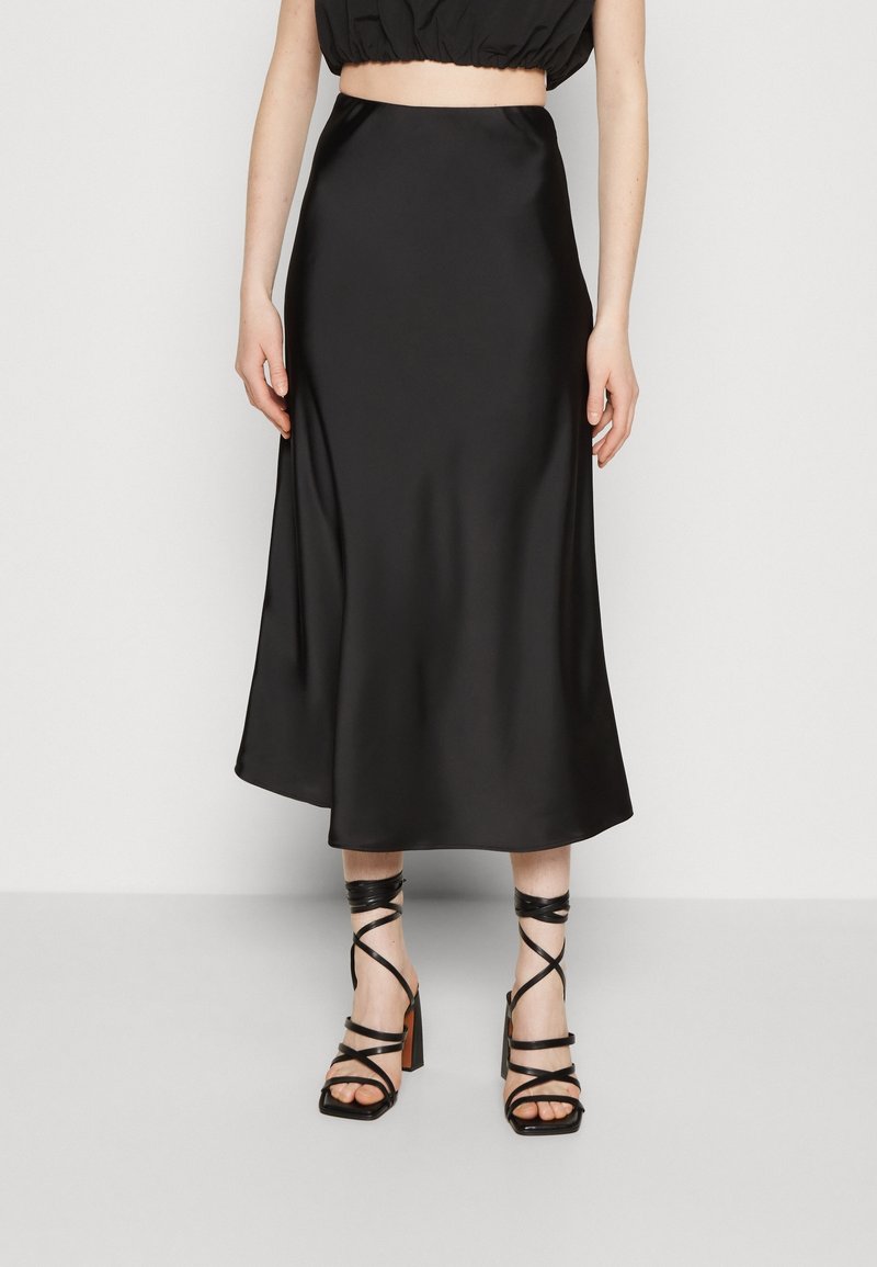 Zwarte satijnen midi-rok met een gladde textuur, uitlopende zoom en hoge taille, gecombineerd met zwart, gespschoenen met hoge hakken.