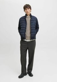Doudoune bleu marine foncé avec poignets côtelés, portée sur un pull en maille beige texturé, associée à un pantalon de costume foncé et des chaussures noires.