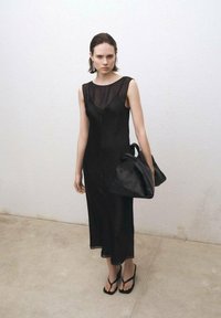 Schwarzes ärmelloses Kleid mit einer transparenten oberen Schicht, lockerer Passform und dezentem V-Ausschnitt. Ergänzt durch eine schwarze lässige Tasche und Flip-Flop-Sandalen.