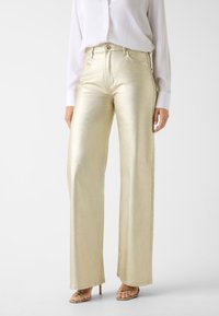 Metalen gouden wijde pijpen broek met een gladde textuur, voorzien van een tailleband met knoopsluiting en zijzakken. Gestyled met een witte blouse.