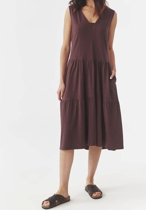 Midi šaty bez rukávů v tmavě hnědé barvě s výstřihem do V a vícevrstvou sukní, nošené s černými sandály s překříženými pásky.