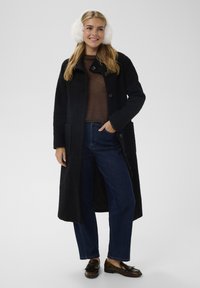 Cappotto lungo nero con finitura testurizzata, davanti con bottoni e tasche laterali. Abbinato a un maglione marrone, jeans blu e mocassini marroni. Paraorecchie in pelliccia bianca.