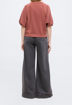 Personne portant un haut à manches courtes couleur rouille avec des manches bouffantes et un pantalon large gris foncé, debout de dos sur un fond uni.