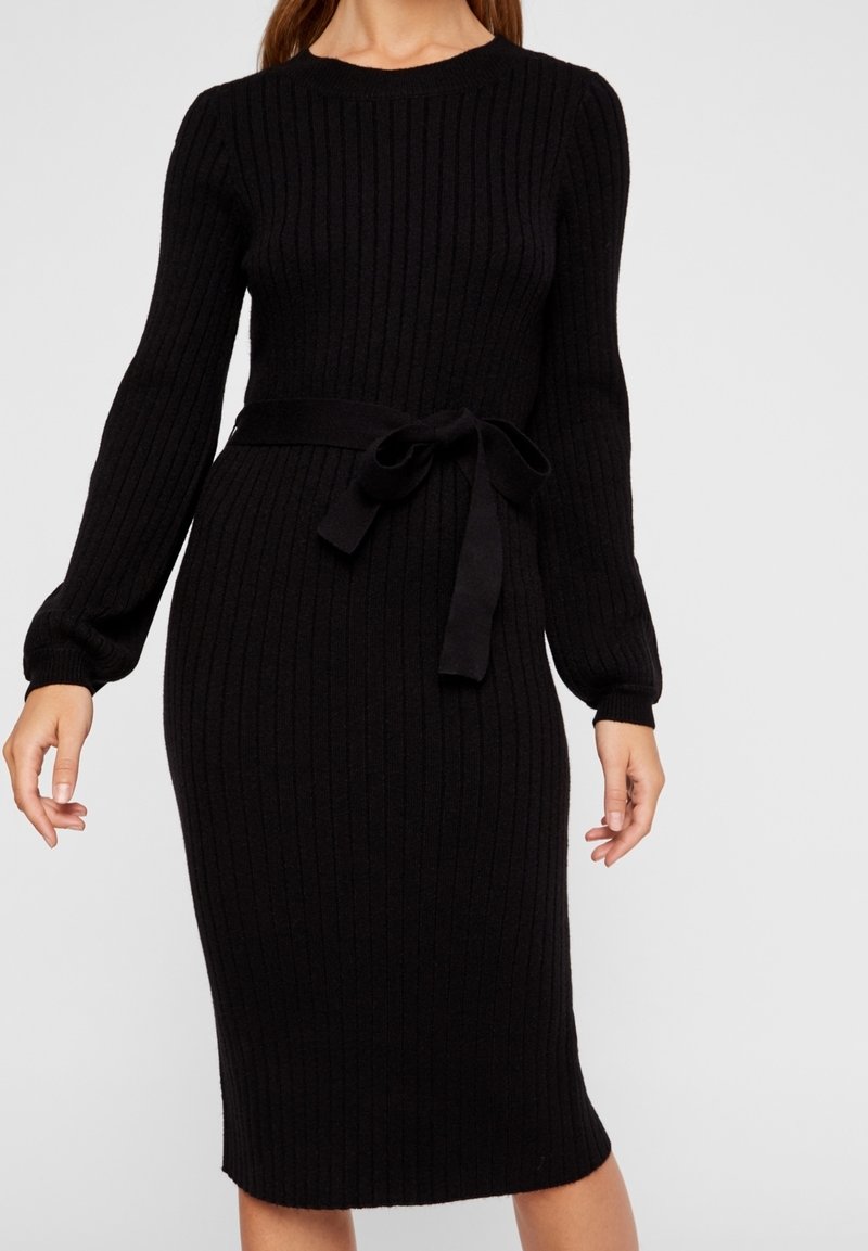 Femme portant une robe midi en tricot côtelé noire ajustée avec manches longues et ceinture en tissu nouée à la taille, debout devant un fond uni.