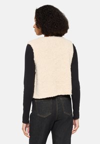 Cremfarbener Fleece-Weste mit texturiertem Finish, kurzer Passform und offenem Rücken, über einem schwarzen Langarmshirt getragen, kombiniert mit dunklen Denim-Hosen.