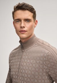 Beige Zip-Jacke mit strukturiertem Blumenmuster, geripptem Kragen und Schulterakzenten, ausgestattet mit einer glatten Stoffoberfläche.