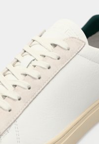 White leather sneaker with a suede beige accent. Features flat cream laces and a rubber sole. Embossed logo on the tongue.