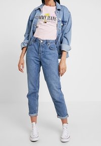 Chaqueta de mezclilla azul claro sobre una camiseta rosa con "TOMMY JEANS" estampada, combinada con jeans azul de tiro alto y zapatillas blancas.