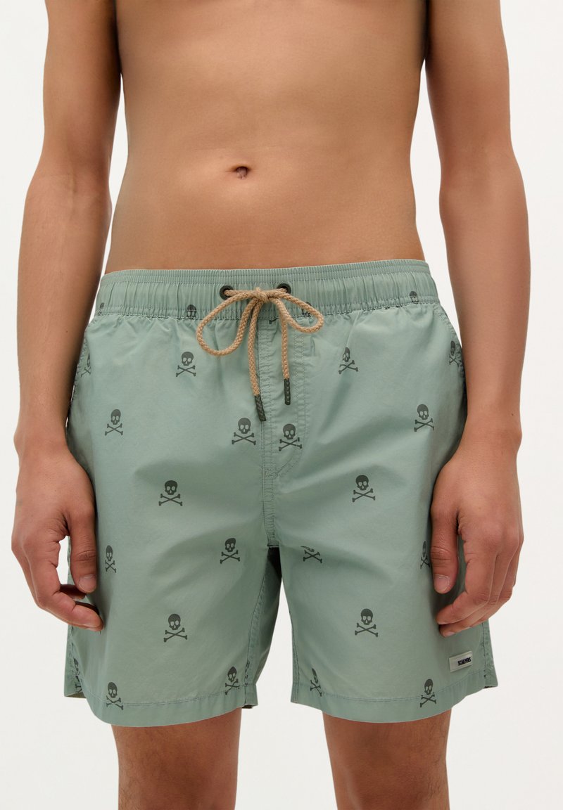 Shorts de bain vert clair avec un motif de skull et os croisés, dotés d'une taille élastique et d'un cordon de serrage. Texture lisse, longueur jusqu'aux genoux.
