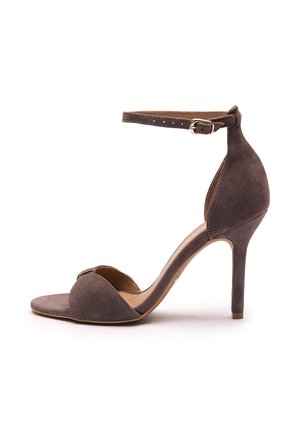 Sandalia de tacón stiletto de ante marrón con correa en el tobillo y hebilla dorada, puntera abierta y tacón alto y delgado sobre fondo blanco.
