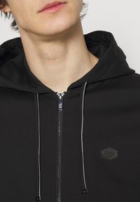 Felpa con zip nera dal finish testurizzato, dotata di chiusura frontale con zip, cordoncini e un logo esagonale sul petto.