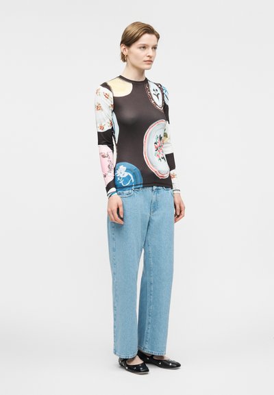 Γυναίκα που φορά πολύχρωμο μακρυμάνικο τοπ με λουλουδάτα σχέδια, ανοιχτό μπλε τζιν με φαρδιά πόδια και μαύρα επίπεδα παπούτσια με διακοσμητικά στοιχεία.