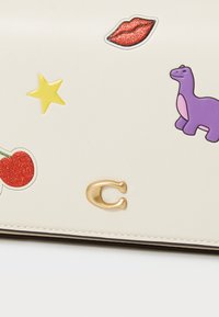 Cream-colored leather purse featuring colorful stickers: red lips, a purple dinosaur, yellow star, and red cherries. Gold logo accent.