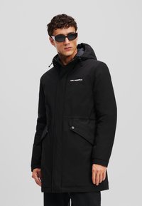 Sort polstret parka med hætte, der har en lynlåslukning, to frontlommer og et logo på brystet. Glat tekstur, længde til midt på låret.