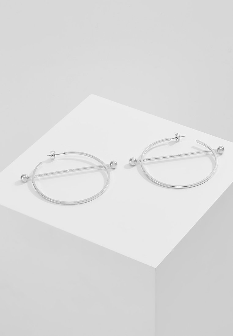 Boucles d’oreilles créoles minimalistes en argent avec une barre droite traversant chaque cercle, reposant sur une surface géométrique blanche sur un fond neutre.