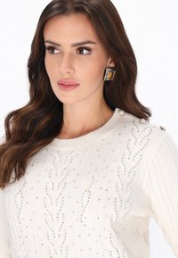 Witte gebreide trui met kabelbreisel en iriserende kraaldetails, met decoratieve knopen op de schouder en parelaccenten.