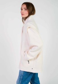 Lichtroze fleecejack met een hoge kraag, oversized pasvorm, zijzakken en ritsdetails, gecombineerd met blauwe denim jeans.