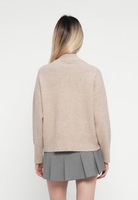 Maglione beige lavorato a maglia con collo alto e spalle scese, abbinato a una gonna grigia a pieghe, visto da dietro.