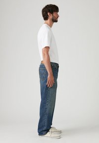 Bela bombažna majica, modre jeans hlače z izpranim videzom, stranski pogled, priložnostna prilagoditev, bele čevlje z gumijastim podplatom, enobarvno ozadje.