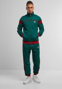 Zielony i czerwony dres wykonany z miękkiego materiału, składający się z kurtki na zamek z logo oraz dopasowanych spodni z bocznymi paskami. Noszony z białymi sneakerami.